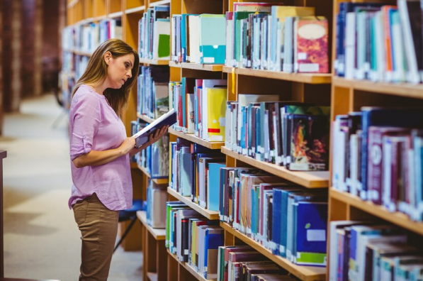 Freepik | Una mujer sostiene un libro abierto en sus manos, mientras lo revisa frente a una estantería dentro de una biblioteca.